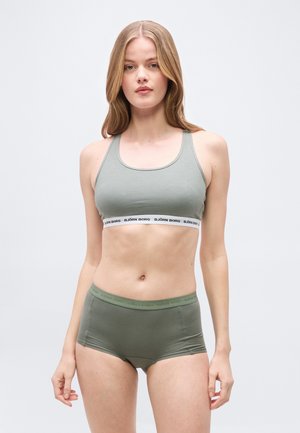 Νεαρή γυναίκα με μακρύ ξανθό μαλλί, φοράει ανοιχτόγκρι αθλητικό σουτιέν και αντίστοιχα πράσινα σορτς με ψηλή μέση, μπροστά σε απλό φόντο.