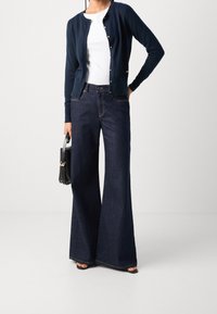 Námořnický svetr s stříbrnými knoflíky přes bílou blůzu, doplněný širokými tmavými džíny. Modelka drží černou kabelku.