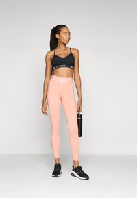 Black sports bra with Nike logo, pink leggings with "NIKE" at waistband, black sneakers, holding a dark water bottle. Smooth, fitted material.