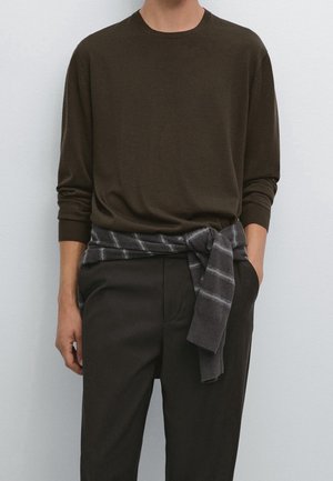 Person iført en mørkebrun langærmet skjorte og matchende bukser med en gråstribet sweater bundet om livet.