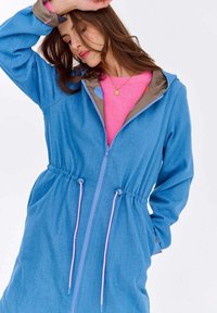 Kobieta ubrana w niebieską kurtkę z zamkiem i różowymi sznurkami na kapturek, w jasnoróżowym swetrze, przechylająca głowę, z uniesioną jedną ręką na białym tle.