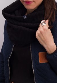 Black knitted infinity scarf draped around the neck, paired with a navy quilted jacket featuring a leather patch and silver zipper detailing.