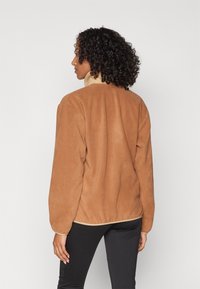 Brun konstläderjacka med en beige krage, långa ärmar och elastiska manschetter, bärs över svarta byxor. Mjuk textur och avslappnad passform.