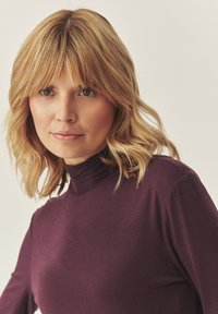 Lila, magasnyakú felső sima anyagból. A modell hullámos, válltól hosszú szőke hajjal és természetes sminkkel rendelkezik.