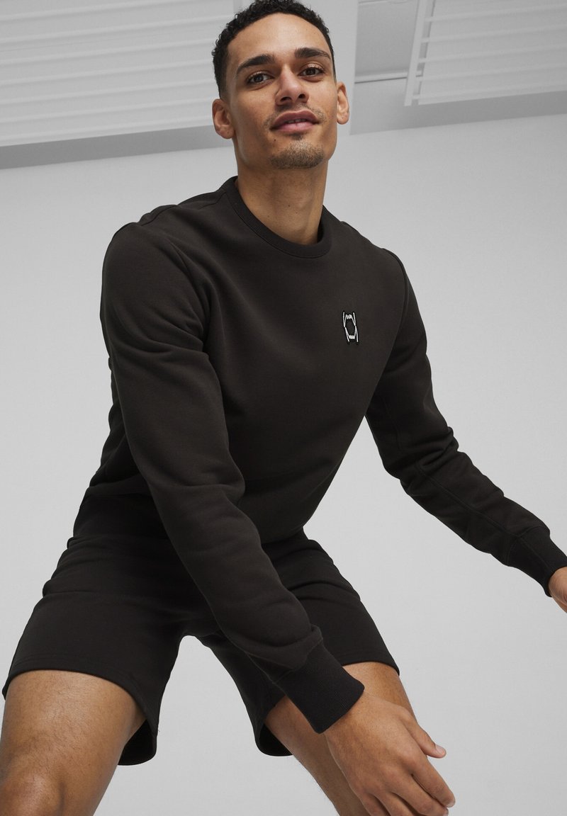 Felpa nera con colletto e polsini a costine, caratterizzata da un piccolo logo bianco sul petto. Indossata con pantaloni corti neri abbinati, che mettono in risalto un look sportivo.