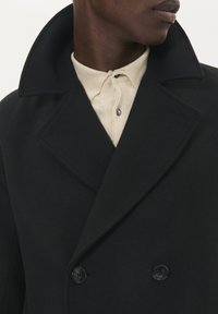 Čierny vlnený kabát s vysokým golierom a dvojradovým zapínaním. Má veľké, okrúhle čierne gombíky a mäkkú textúru. Pod ním je béžová košeľa.