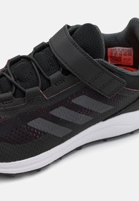 Chaussure de sport noire avec tige en mesh, semelle en caoutchouc, bande Velcro et trois rayures grises distinctives sur le côté. Accents rouges.
