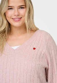 Svetlo roza pletena jopica z V-izrezom, podrobno pletenim vzorcem in majhnim rdečim vezenim srcem na prsih. Mehak občutek.