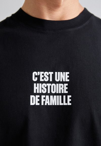 Sort t-shirt i sort bomuld med hvid tekst, der står "C’EST UNE HISTOIRE DE FAMILLE" i fed, stablet typografi. Rund hals.