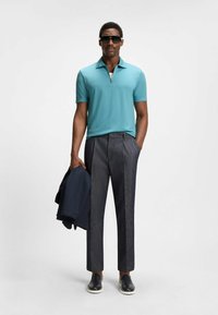 Jasnoniebieska koszulka polo na zamek, szare wzorzyste spodnie, czarne trampki, trzymając granatową kurtkę. Okulary przeciwsłoneczne dopełniają wygląd.