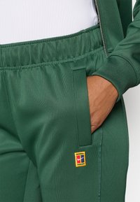 Pantaloni sportivi verdi realizzati in poliestere liscio, con vita elastica, tasca laterale e una toppa con logo multicolore sulla coscia.