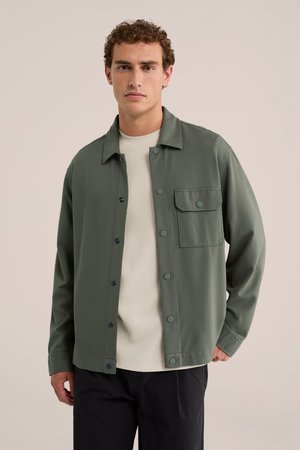Jeune homme aux cheveux bouclés portant une veste verte boutonnée sur une chemise blanche texturée et un pantalon noir, debout devant un fond uni.