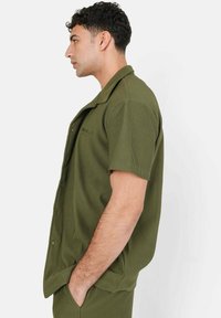 Korte mouwen, olijfgroen knoopshirt van ribgebreide stof met een ontspannen pasvorm. Heeft een kraag en een voorzakdetail.