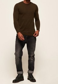 Chemise en maille vert foncé à manches longues associée à un jean en denim noir retroussé et des bottes noires à lacets, mettant en valeur une silhouette ajustée et un tissu texturé.