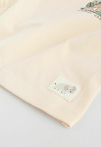 T-shirta w kolorze brzoskwiniowym, wykonana z bawełny o miękkiej teksturze. Posiada małą metkę z tkaniny przedstawiającą minimalistyczny wzór słonia.