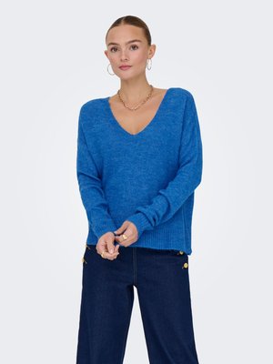 Mujer que lleva un suéter azul con cuello en V, pantalones anchos de pierna oscura con botones dorados, pendientes de aro y un collar de cadena dorada, de pie sobre un fondo blanco.