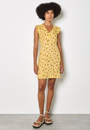 Femme debout portant une robe sans manches jaune à fleurs avec des épaules à volants et des sandales compensées beiges, posant devant un mur blanc uni.