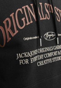 Sudadera negra con letras en relieve en marrón que dicen "ORIGINALS" y texto en blanco. Cuenta con cordones con punta de plata y una tela suave y texturizada.