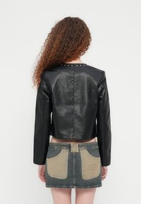 Μαύρο δερμάτινο κοντό μπουφάν με μακριά μανίκια, διακοσμημένο με απλό σχέδιο στην πλάτη και ασημένιες τρουκς κατά μήκος του λαιμού. Φορεμένο με τζιν φούστα.