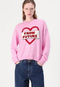 Pullover rosa con collo a giro, caratterizzato da un design a forma di cuore rosso e la scritta "FROM FUTURE" in lettere in grassetto, abbinato a jeans blu scuro.