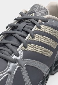 Κοντινή λήψη γκρι αθλητικού παπουτσιού Adidas με υφασμένες μπεζ ρίγες και λεπτομερή κορδόνια σε λευκό φόντο.