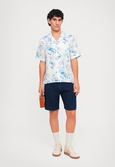 Uomo in piedi che indossa una camicia bianca a maniche corte con stampe blu marine, pantaloni corti blu scuro, calze bianche, scarpe da ginnastica beige, mentre tiene in mano una piccola scatola marrone.