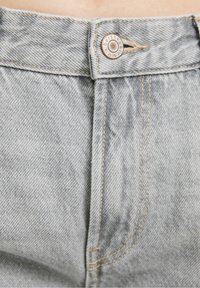 Jeans de mezclilla gris claro con cierre de botón y costuras naranjas. Presentan un logo en el botón y una textura suave.