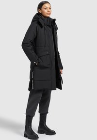 Black padded parka with a hood, front pockets, and zip accents. Features a sleek design and tall black boots. Smooth texture and warm material.