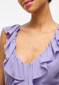 Svetlo-lavanda bluza z naborki, z globokim V-izrezom, mehkim materialom in zbranimi detajli ob ramenih.