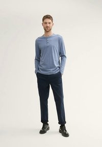 Helehall sinine pikkade varrukatega henley-särgi, millel on kolm nööpi, kombineeritud mereväe värvi pükste ja mustade pits-up kingadega, seisab neutraalse tausta ees.