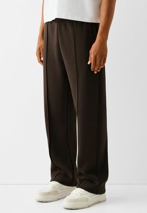 Mann trägt lockere schwarze Hose mit vorderen Nahtdetails und weißen Sneakern, steht vor einem einfarbigen hellgrauen Hintergrund.