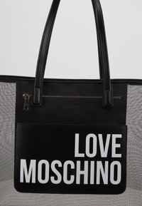 Svart tote-väska med konstläderdetaljer, har en ficka med dragkedja, mesh-sidor och stor vit text som säger "LOVE MOSCHINO" på framsidan.