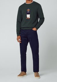Sudadera verde oscura con un estampado gráfico de un personaje, combinada con chinos azul marino y zapatillas color canela. Ajuste cómodo y diseño casual.
