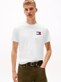 White cotton t-shirt featuring a small, horizontal logo patch in red, blue, and white. Paired with olive green pants and a black belt.