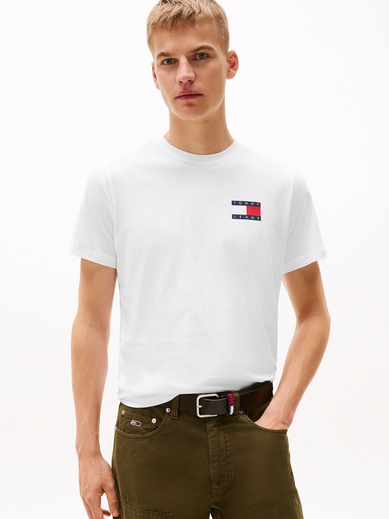White cotton t-shirt featuring a small, horizontal logo patch in red, blue, and white. Paired with olive green pants and a black belt.