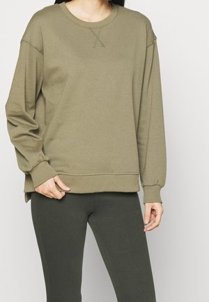 Kvinde iført olivengrøn langærmet sweatshirt og mørkegrønne tætsiddende bukser, stående med den ene hånd ved taljen og den anden afslappet.