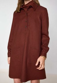Abito color ruggine con colletto, apertura a bottoni frontale, maniche lunghe e due tasche frontali. Tessuto liscio con una vestibilità leggermente rilassata.