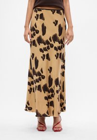 Beige maxi-jurk met donkere, abstracte vlekken, gedragen met een zwarte top en rode sandalen met hoge hakken.