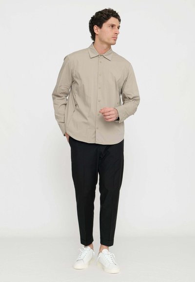 Camicia button-up beige con colletto e maniche lunghe, abbinata a pantaloni neri affusolati e sneakers bianche. Tessuto liscio, design minimalista.