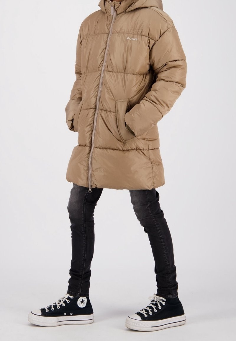 Doudoune couleur camel avec capuche et fermeture éclair frontale, ornée de panneaux cousus. Portée avec un jean skinny noir et des baskets Converse noires.