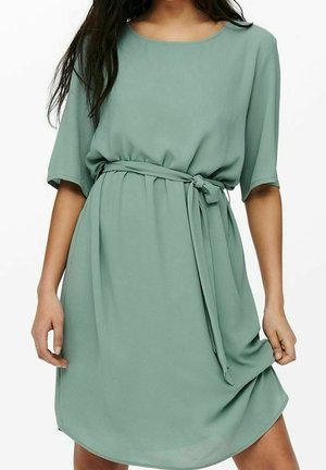 Woman wearing a loose, sage green knee-length dress with elbow-length sleeves and a tied waist belt, holding the hem with one hand.