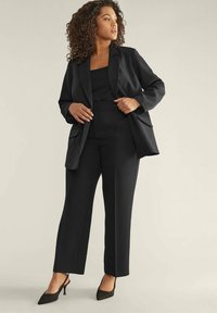 Completo nero su misura con un blazer morbido dotato di tasche, pantaloni dritti e una blusa elegante. Il tessuto appare liscio con una finitura opaca.