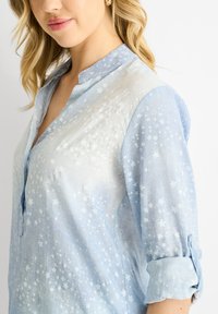 Blusa azzurro chiaro con motivo a stelle, dotata di colletto e maniche arrotolate, realizzata in un tessuto morbido e texturizzato.