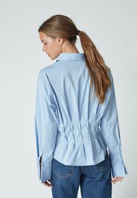 Light blue shirt with a fitted waist and pleated back. Long sleeves feature slit cuffs, tailored collar, and smooth fabric texture.