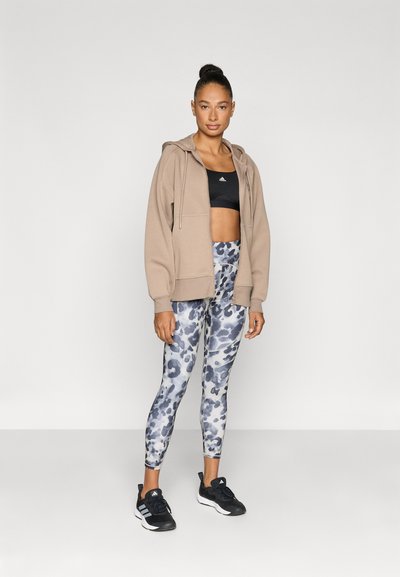 Felpa con cappuccio beige con zip sopra un reggiseno sportivo nero, abbinata a leggings blu e grigi con motivo, e scarpe da ginnastica nere.