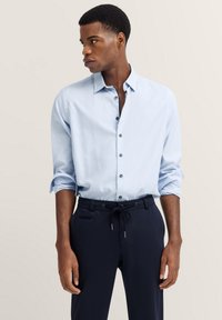 Hellblaues Button-up-Hemd aus Baumwolle mit einem schmalen Kragen, langen Ärmeln und dunklen Knöpfen. Kombiniert mit navyblauen Hosen.