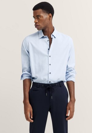Light blue button-up shirt in cotton fabric, featuring a narrow collar, long sleeves, and dark buttons. Paired with navy pants.