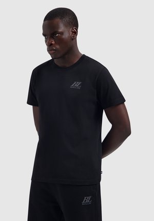 Mann trägt schwarzes Ballin-T-Shirt und passende Shorts vor schlichtem hellem Hintergrund, Hände hinter dem Rücken.