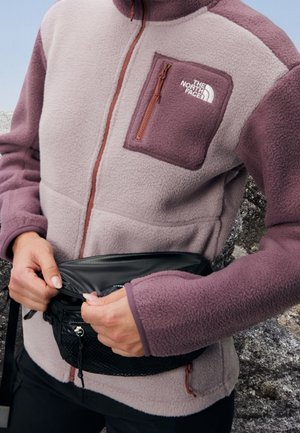 Personne portant une veste polaire bicolore violette avec le logo The North Face, ouvrant une sacoche noire à la taille en plein air près d'un terrain rocheux.