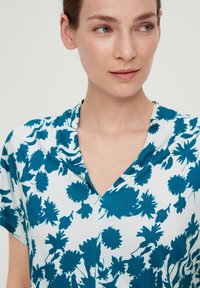 V-hals top met een teal bloemenpatroon op een witte achtergrond. Gemaakt van zachte stof met een relaxte pasvorm en korte mouwen.